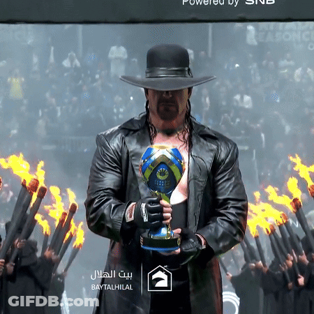A Man In A Hat Holds A Trophy In Front Of Flames And A Sign That Says Baytal Hilal GIF