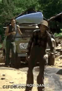 A Man In A Hat Is Dancing In Front Of A Truck With A Kayak On Top Of It . GIF