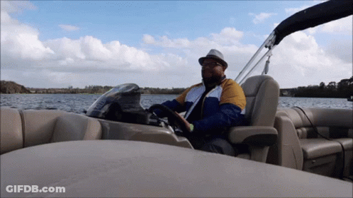 A Man In A Hat Is Driving A Pontoon Boat On A Lake GIF