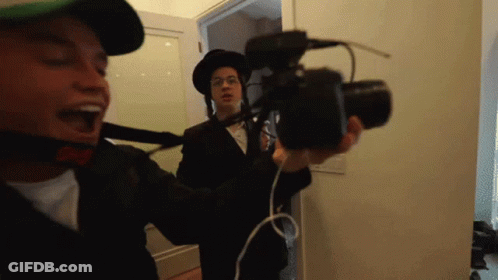 A Man In A Hat Is Holding A Camera While Another Man In A Hat Looks On GIF