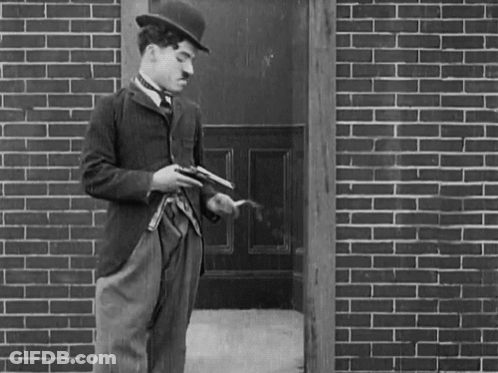 A Man In A Hat Is Holding A Gun In Front Of A Brick Building GIF