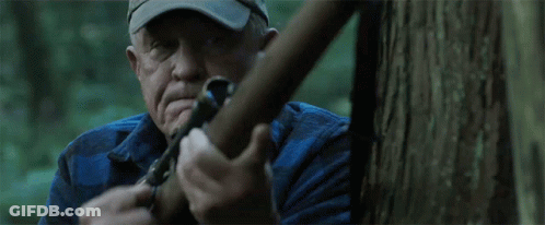 A Man In A Hat Is Holding A Sniper Rifle In Front Of A Tree GIF