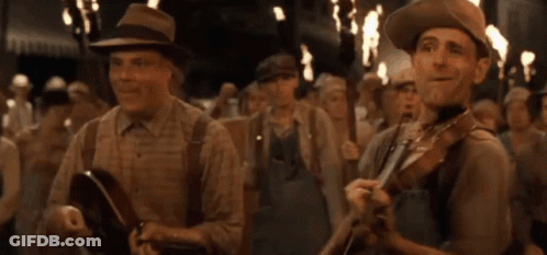A Man In A Hat Is Playing A Violin In Front Of A Crowd Of People GIF