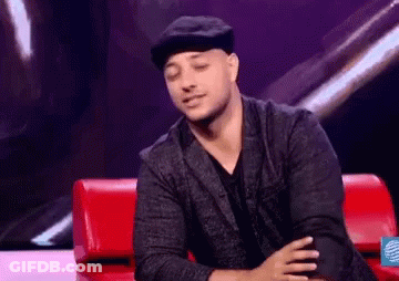 A Man In A Hat Is Sitting On A Red Couch With His Hands Folded . GIF