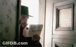 A Man In A Hat Is Standing In Front Of A Door Holding A Piece Of Paper That Says Do N't Say Anything GIF