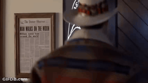 A Man In A Hat Is Standing In Front Of A Framed Newspaper That Says Man Walks On The Moon GIF