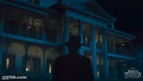 A Man In A Hat Is Standing In Front Of A Haunted Mansion GIF