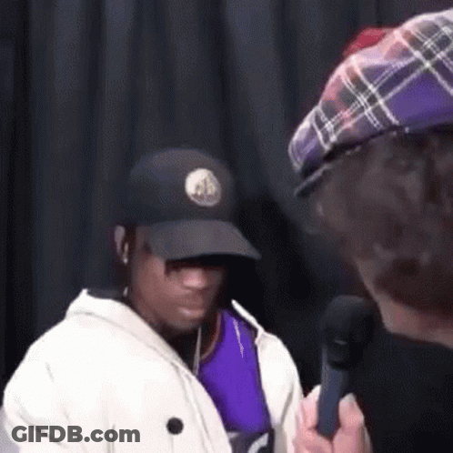 A Man In A Hat Is Talking Into A Microphone While Another Man Looks On . GIF