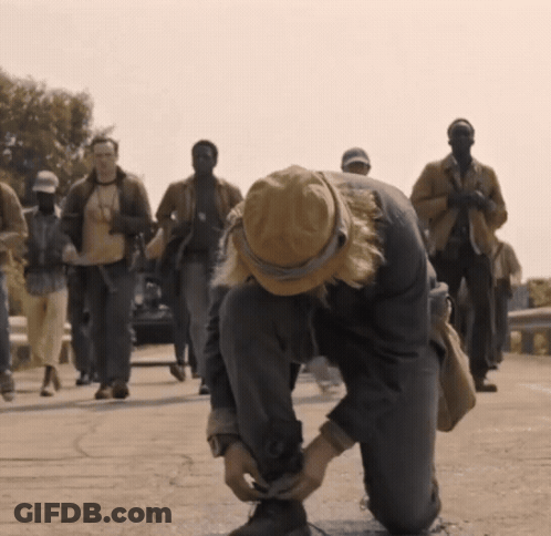 A Man In A Hat Is Tying His Shoelaces While A Group Of People Walk Behind Him GIF