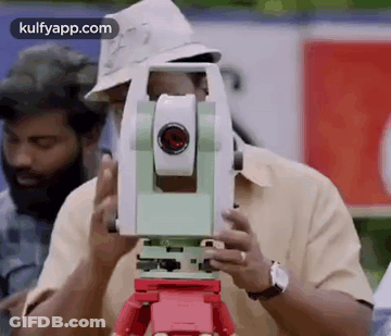 A Man In A Hat Is Using A Theodolite On A Tripod To Measure Something . GIF