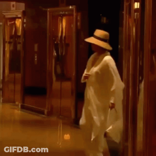 A Man In A Hat Is Walking Down A Hallway Holding A Bag . GIF