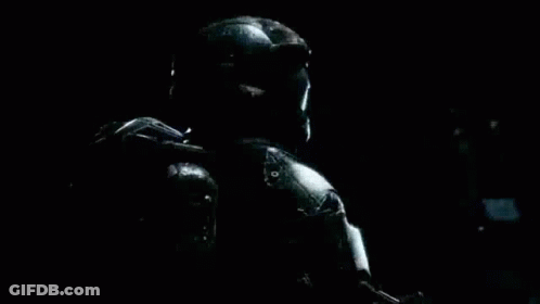 A Man In A Helmet And Armor Is Holding A Gun In A Dark Room . GIF