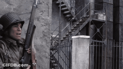 A Man In A Helmet Holds A Gun In Front Of A Building GIF