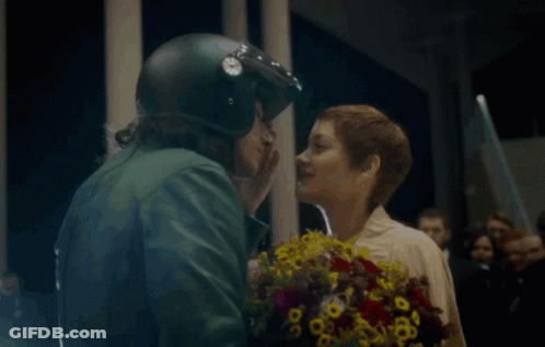 A Man In A Helmet Kisses A Woman Holding Flowers GIF