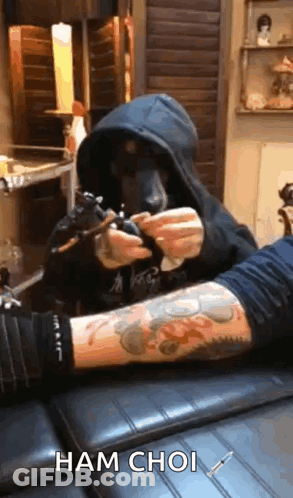 A Man In A Hood Is Getting A Tattoo On Another Man 'S Arm . GIF