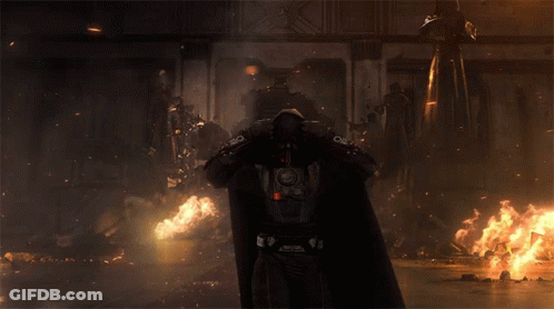 A Man In A Hooded Cape With A Red Light On The Back Stands In Front Of A Fire GIF