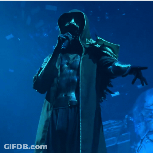 A Man In A Hooded Coat Is Singing Into A Microphone In A Dark Room GIF