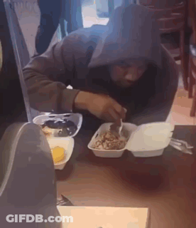 A Man In A Hooded Jacket Is Eating Food From A Styrofoam Container . GIF