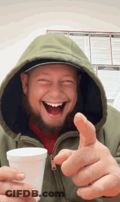 A Man In A Hooded Jacket Is Holding A Cup And Giving A Thumbs Up Sign GIF