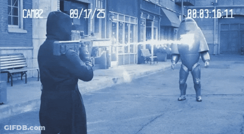 A Man In A Hooded Jacket Is Holding A Gun In Front Of A Camera That Says Cam82 On The Bottom GIF