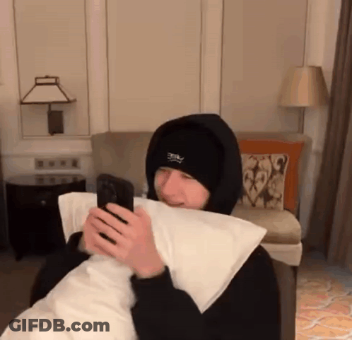 A Man In A Hooded Jacket Is Holding A Pillow And Looking At His Phone . GIF
