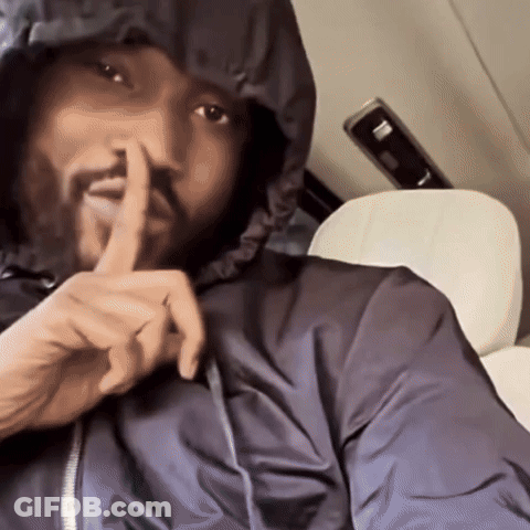A Man In A Hooded Jacket Is Holding His Finger To His Mouth GIF