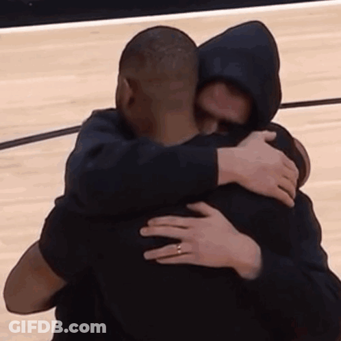 A Man In A Hooded Jacket Is Hugging Another Man On A Basketball Court . GIF