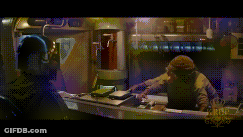 A Man In A Hooded Jacket Is Sitting At A Table In A Dark Room With A Crown On It . GIF
