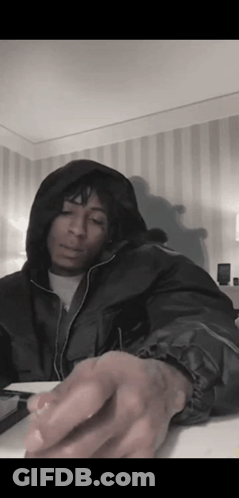A Man In A Hooded Jacket Is Sitting At A Table In A Room With A Striped Wall . GIF
