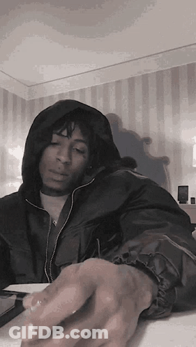 A Man In A Hooded Jacket Is Sitting At A Table With His Hands On The Table . GIF