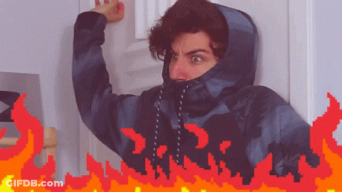 A Man In A Hooded Jacket Is Standing In Front Of A Door With Flames Behind Him GIF
