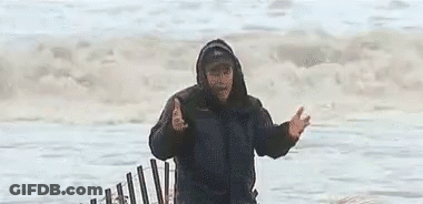 A Man In A Hooded Jacket Is Standing In Front Of A Fence On The Beach . GIF