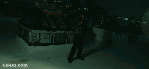 A Man In A Hooded Jacket Stands On A Rooftop Holding An Umbrella And A Microphone GIF