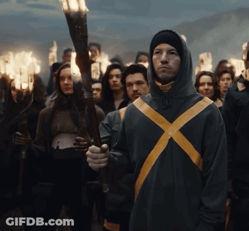 A Man In A Hooded Jacket With A Yellow X On It Holds A Torch In Front Of A Crowd GIF