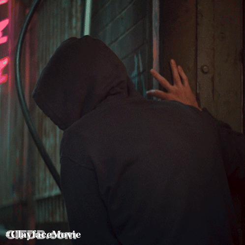 A Man In A Hooded Sweatshirt Is Standing In Front Of A Neon Sign That Says Clayface Movie GIF
