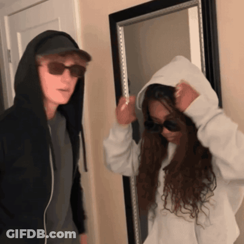 A Man In A Hoodie And Sunglasses Stands Next To A Woman In A Hoodie GIF