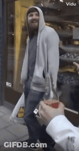 A Man In A Hoodie Holds A Bottle Of Soda In Front Of A Store That Says Vidu On It GIF