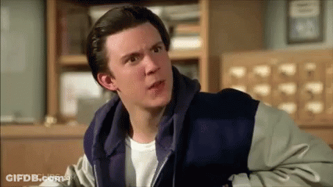 A Man In A Hoodie Is Sitting In Front Of A Bookshelf And Looking At The Camera . GIF
