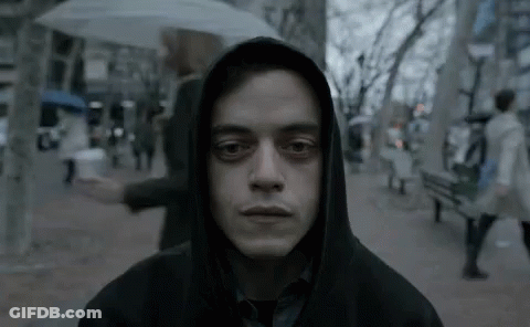 A Man In A Hoodie Is Standing On A Sidewalk With An Umbrella In His Hand . GIF