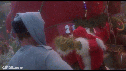 A Man In A Hoodie Talks To A Grinch Who Is Wearing A Santa Hat GIF