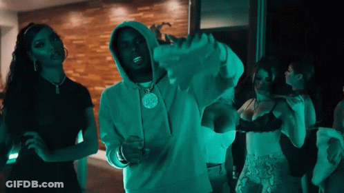 A Man In A Hoodie With A Pendant On His Neck Stands In Front Of Two Women In Bras GIF