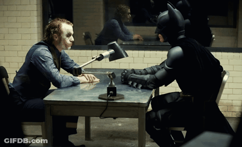 A Man In A Joker Costume Sits At A Table With A Batman Costume GIF