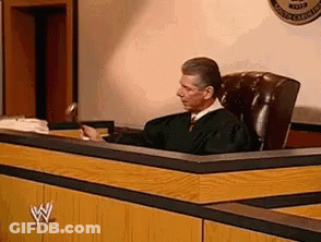 A Man In A Judge 'S Robe Sits At A Desk In A Courtroom . GIF