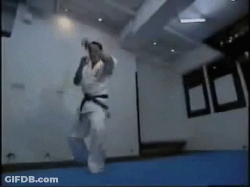 A Man In A Karate Uniform Is Jumping In The Air In A Room . GIF