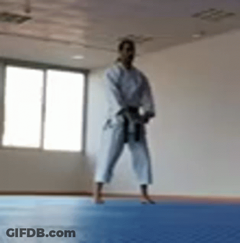 A Man In A Karate Uniform Is Standing On A Blue Mat In A Gym . GIF