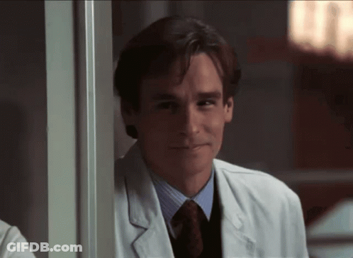 A Man In A Lab Coat And Tie Smiles While Leaning On A Wall GIF