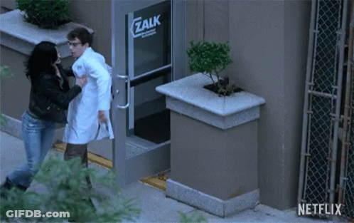 A Man In A Lab Coat Is Being Pushed By A Woman In Front Of A Zalk Building GIF