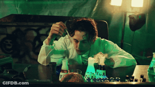 A Man In A Lab Coat Is Sitting At A Table With A Bottle Of Vega On It GIF