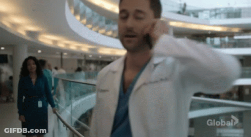 A Man In A Lab Coat Is Talking On A Cell Phone While Walking Down A Hospital Hallway . GIF