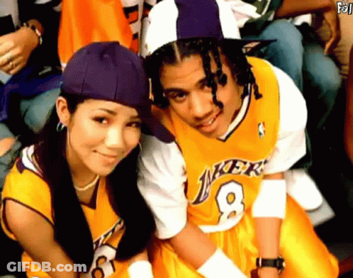 A Man In A Lakers Jersey Sits Next To A Woman Wearing A Purple Hat GIF
